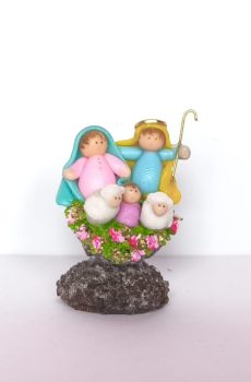 Pesebre artesanal en miniatura con figuras coloridas y ovejas sobre base floral.