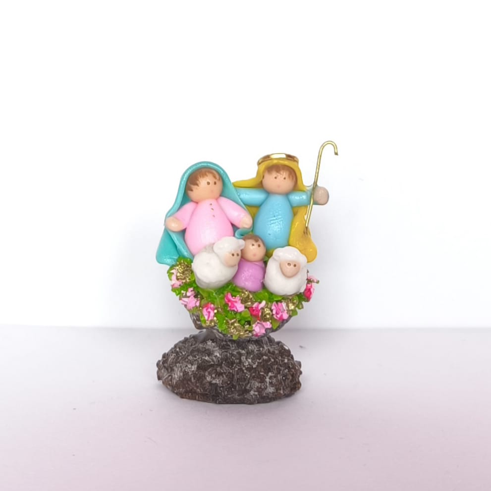 Pesebre artesanal en miniatura con figuras coloridas y ovejas sobre base floral.