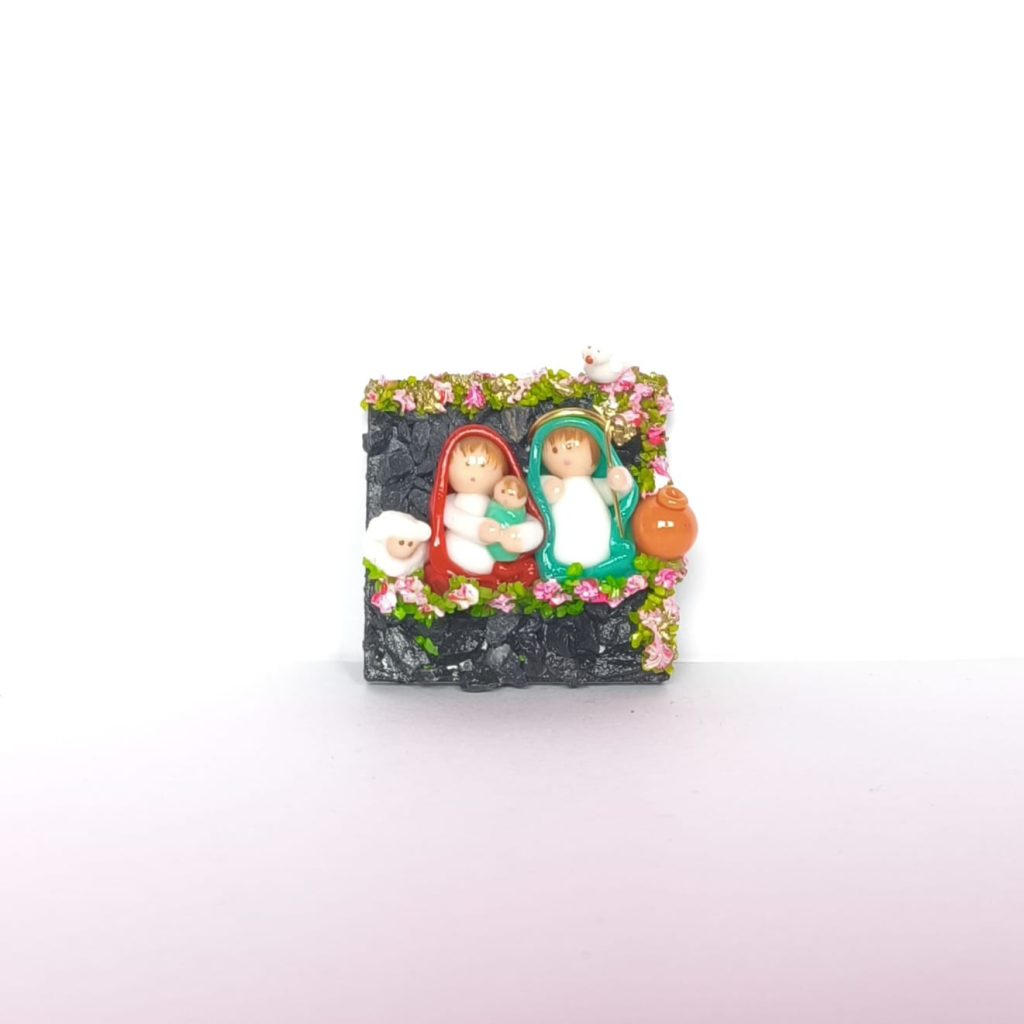 Pesebre en miniatura artesanal con imán, que muestra la Sagrada Familia rodeada de flores y detalles coloridos sobre base oscura.