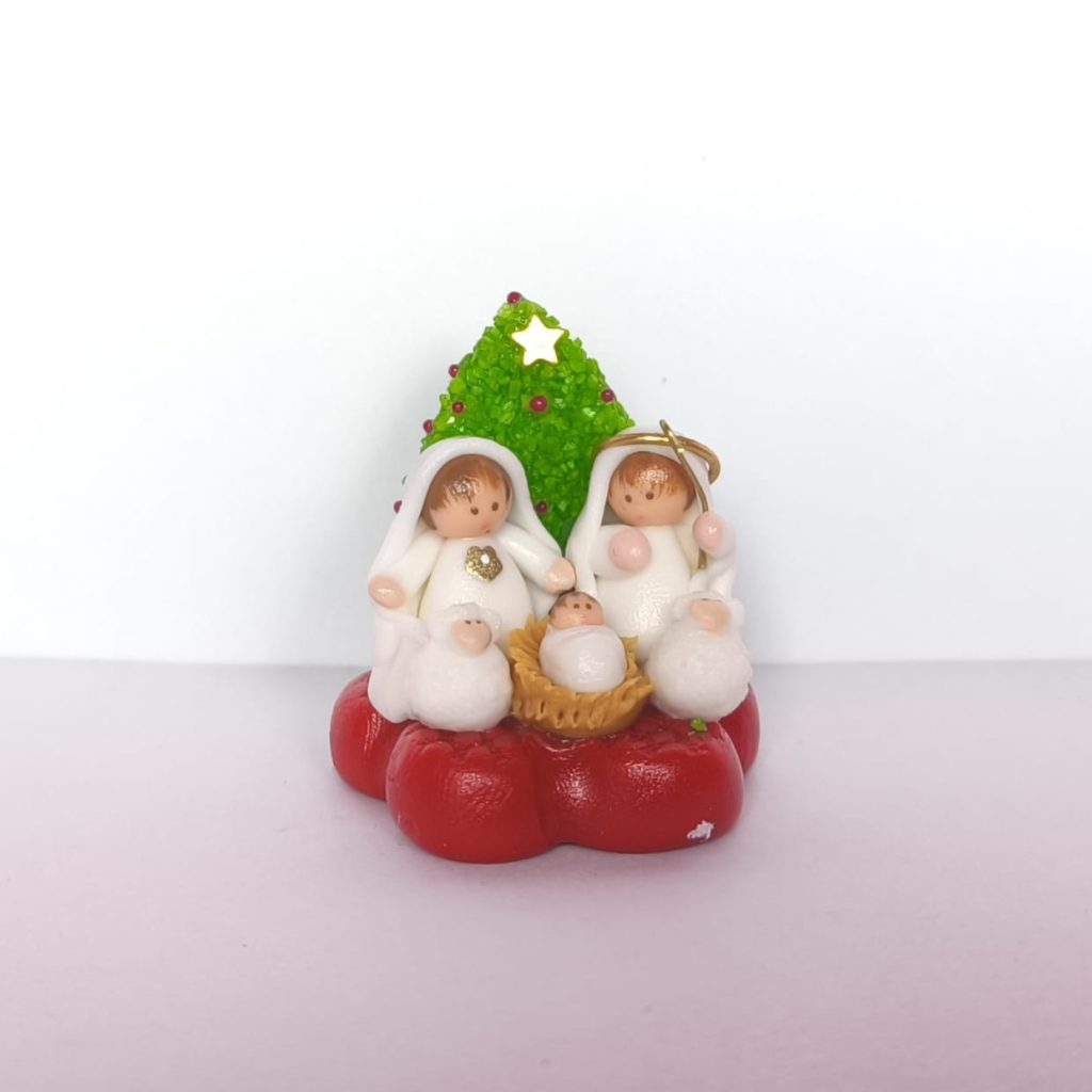 Pesebre en miniatura artesanal con la Sagrada Familia vestida de blanco frente a un árbol de Navidad verde con estrella dorada, sobre base roja.