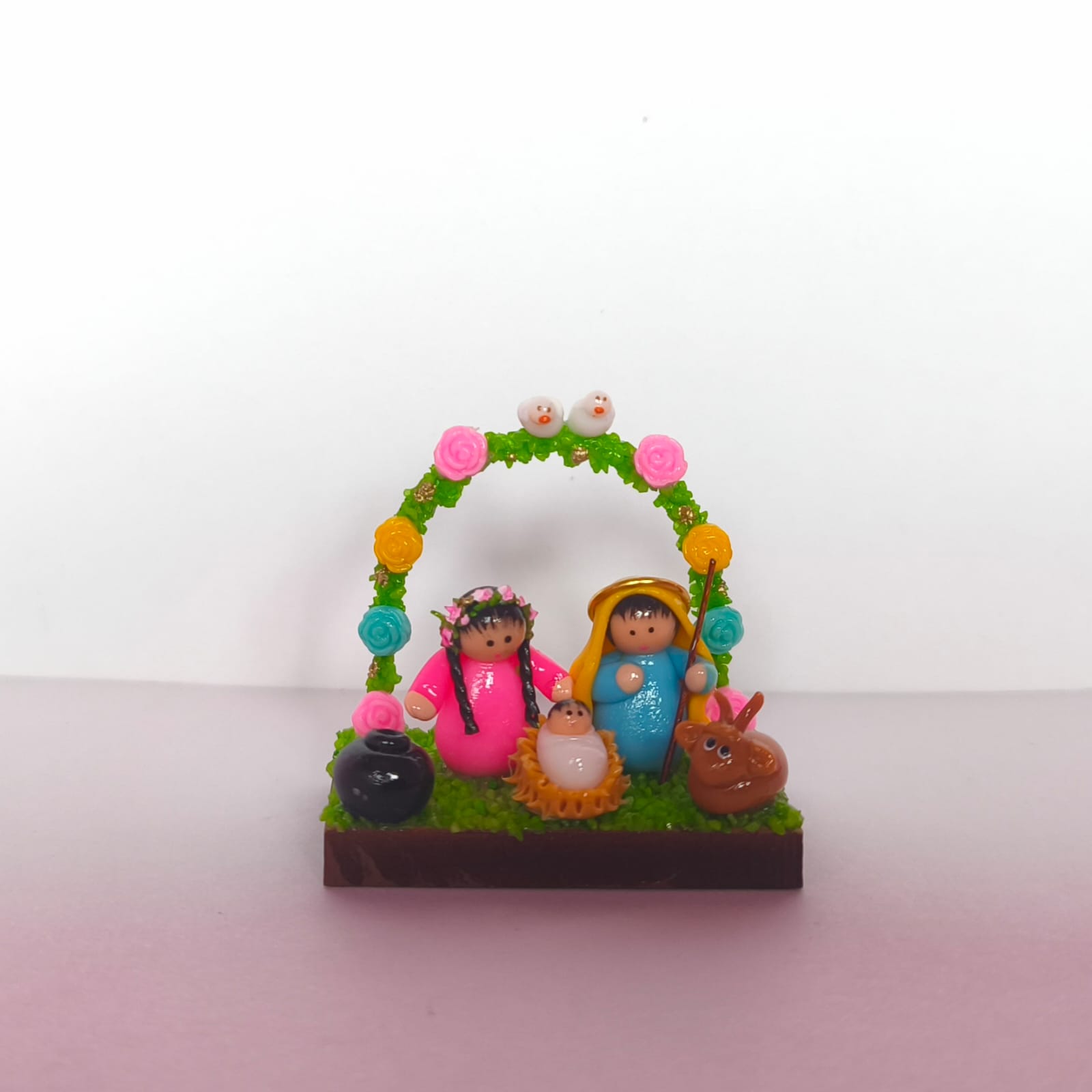 pesebre en miniatura que muestra la Sagrada Familia rodeada con arco de flores y dos palomas sobre base de madera.