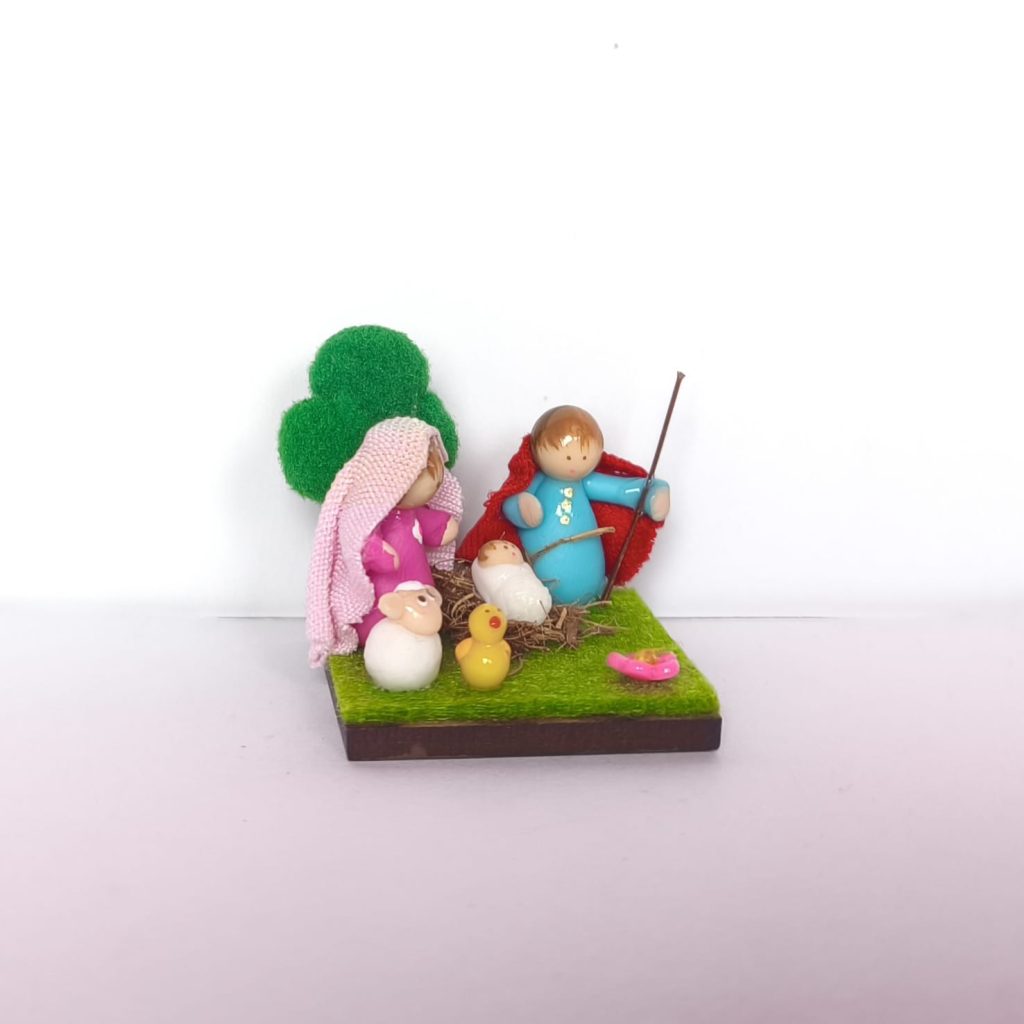 Pesebre en miniatura artesanal con la Sagrada Familia con mantas en tela, sobre base verde con flor y pollito.